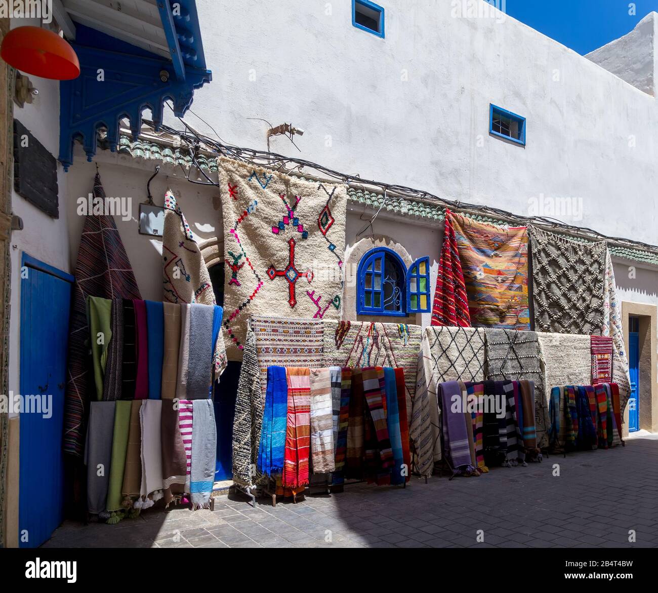 Bunte Stoffe, Teppiche und Homespuns hängen an der typischen weißen und blauen Hauswand, die auf dem Straßenmarkt von Medina.Essaouira, Marokko zu verkaufen ist. Stockfoto