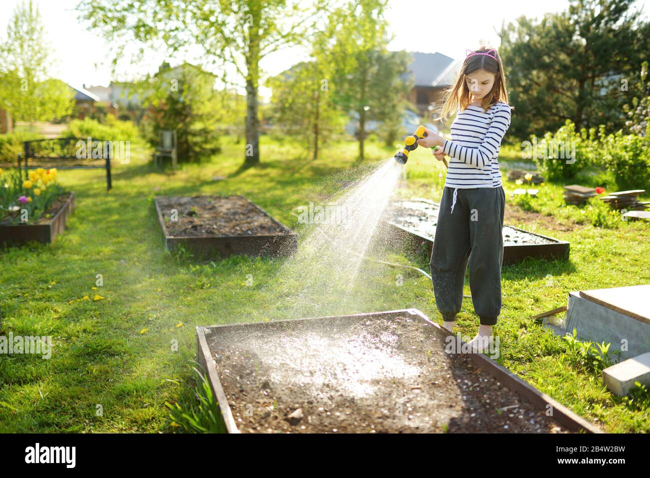 Süße junge Mädchen, die am Sommertag Blumenbeete im Garten gießen. Kind mit Gartenschlauch am sonnigen Tag. Mummys kleiner Helfer. Stockfoto