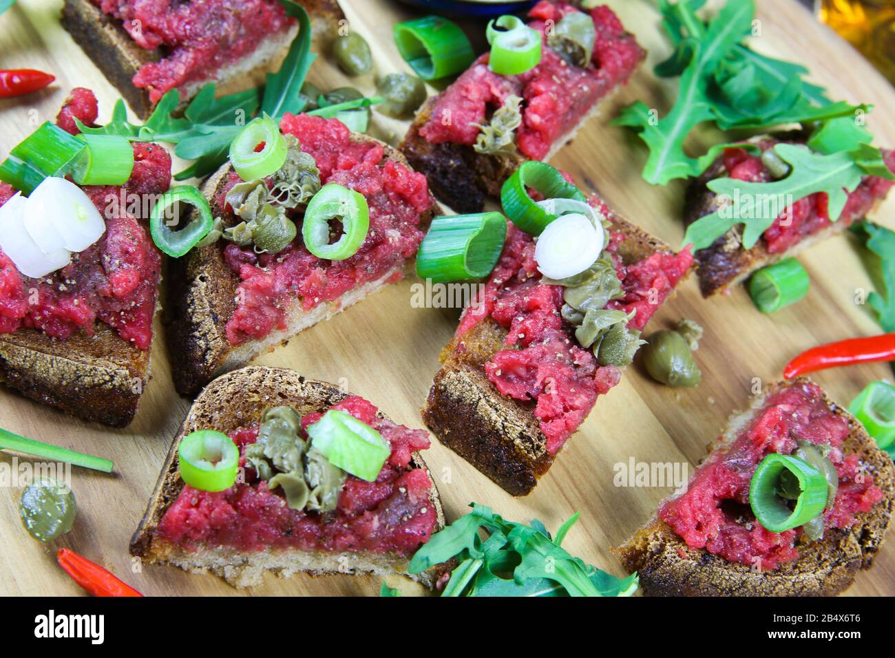 Herzhaft heftiger deutscher Imbiss: Isolierte Holzplatte mit geröstetem tataren-knoblauchbrot, Frühlingszwiebeln, Kapern und Rucola Salatblättern Stockfoto