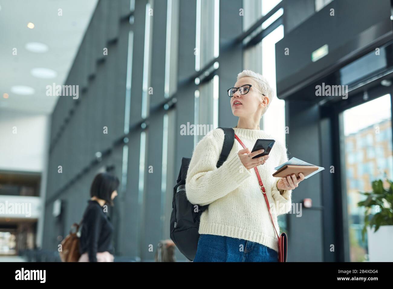 Horizontales Tiefwinkelporträt stilvoller kaukasischer Frauen mit kurzen blonden Haaren, die im Flughafen stehen und Smartphone und Bordkarte fernhalten, Stockfoto