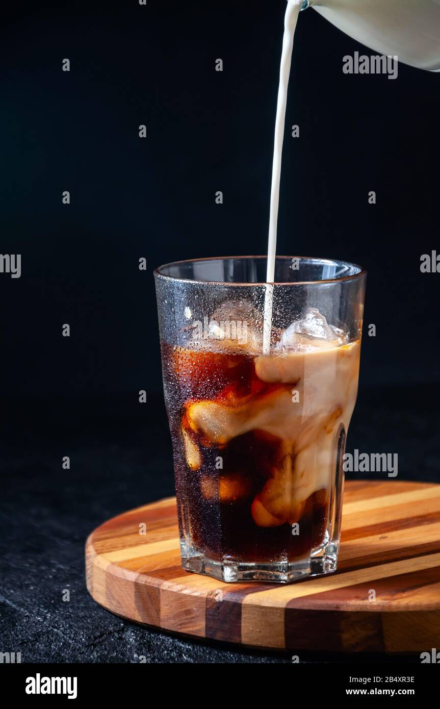 Milch, Die in Eiskaffee in Hochgläserem Glas auf dunklem Hintergrund Gegossen wird. Konzept Erfrischendes Sommergetränk. Stockfoto