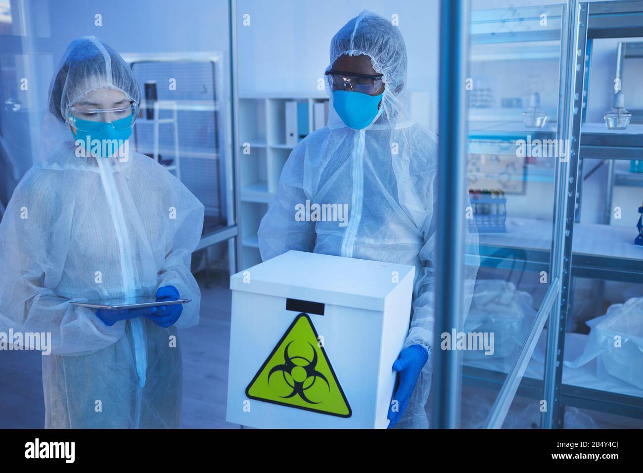 Horizontaler Hochwinkel-Wissenschaftler mit unerkennbarem männlichen Mediakl in schützender Arbeitskleidung bringt Box mit biologischen Gefahren in den Laborraum Stockfoto