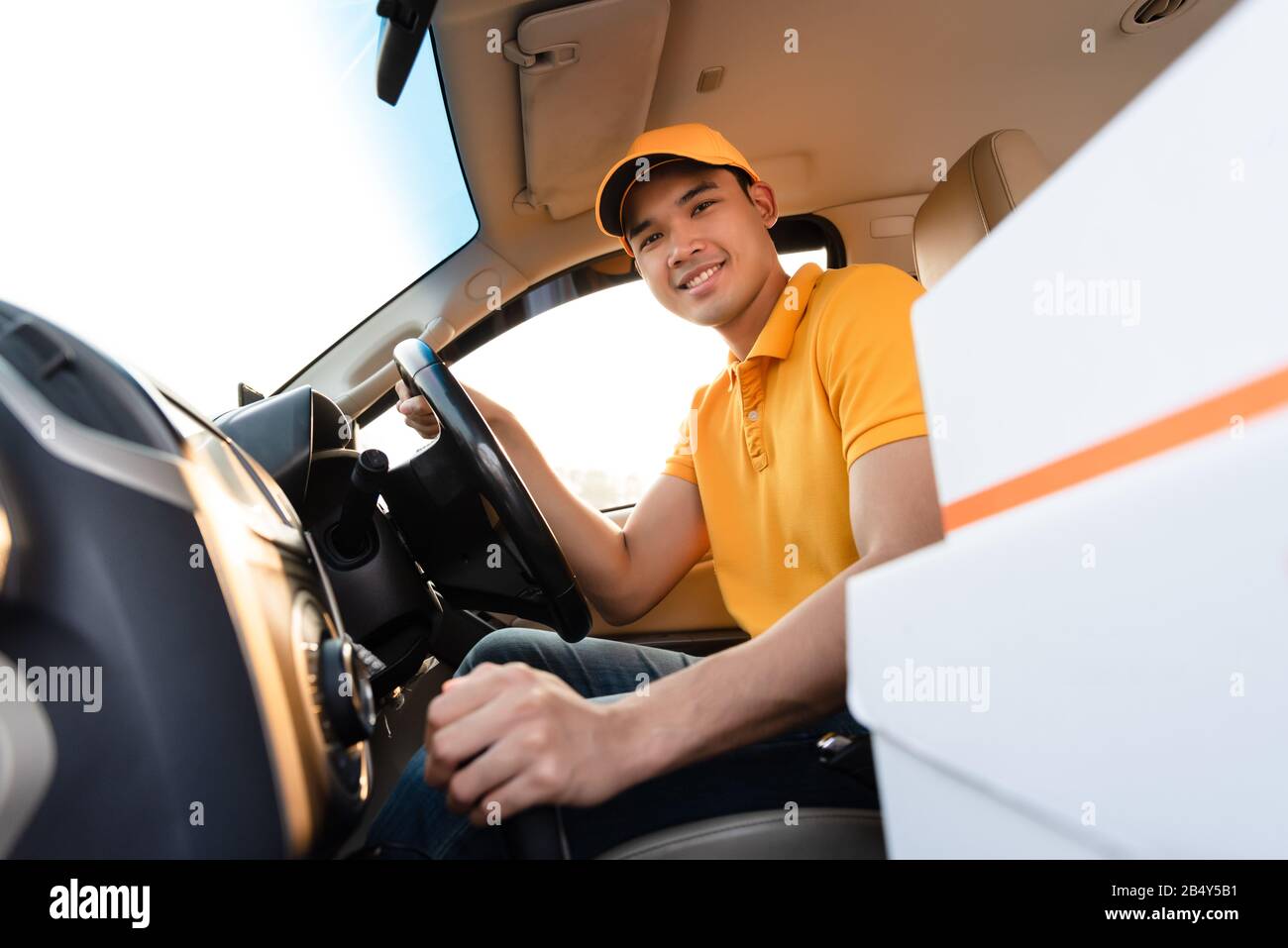 Glücklicher Liefermann in gelber Poloshirt-Uniform mit Paketkarton im Auto. Lächelnder Mann der Kurierzustellung, der im Auto sitzt und den Wahnvorstellung überprüft Stockfoto