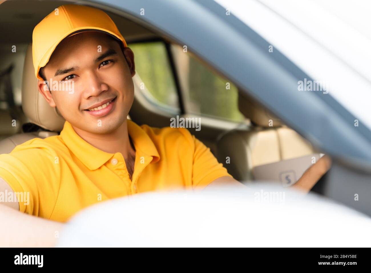 Glücklicher Liefermann in gelber Poloshirt-Uniform mit Paketkarton im Auto. Lächelnder Mann der Kurierzustellung, der im Auto sitzt und den Wahnvorstellung überprüft Stockfoto