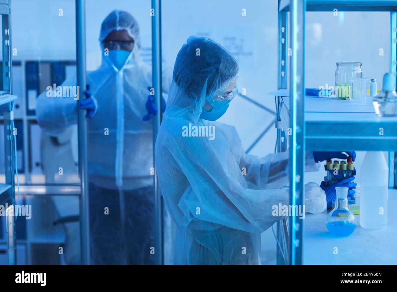 Nicht erkennbare männliche Medizinwissenschaftlerin in schützender Arbeitskleidung, die Tür zum Laborraum öffnet, seine Kollegin, die Reagenzgläser vom Regal nimmt Stockfoto