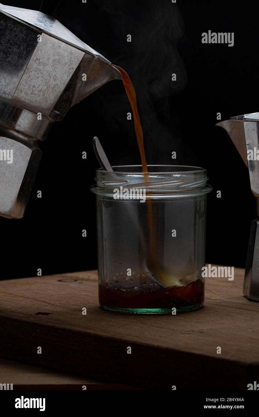 Heißes Dampfgaren erfrischender Kaffee in einem Glas auf dunklem Hintergrund. Gießen von Moka-Hersteller in leeres Glas mit Löffel Stockfoto