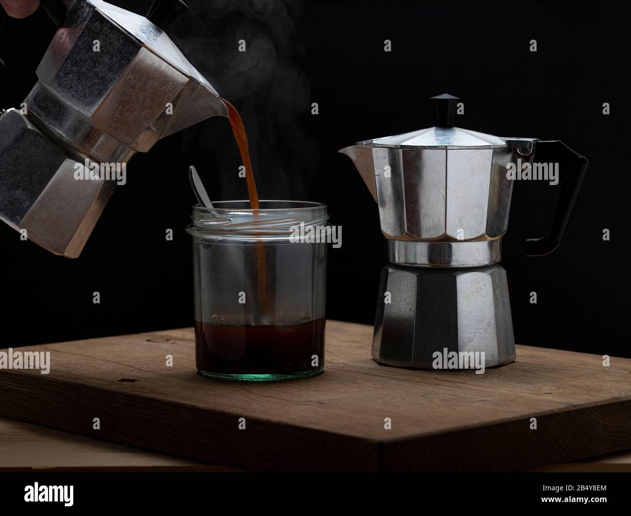 Heißes Dampfgaren erfrischender Kaffee in einem Glas auf dunklem Hintergrund. Gießen von Moka-Hersteller in leeres Glas mit Löffel Stockfoto