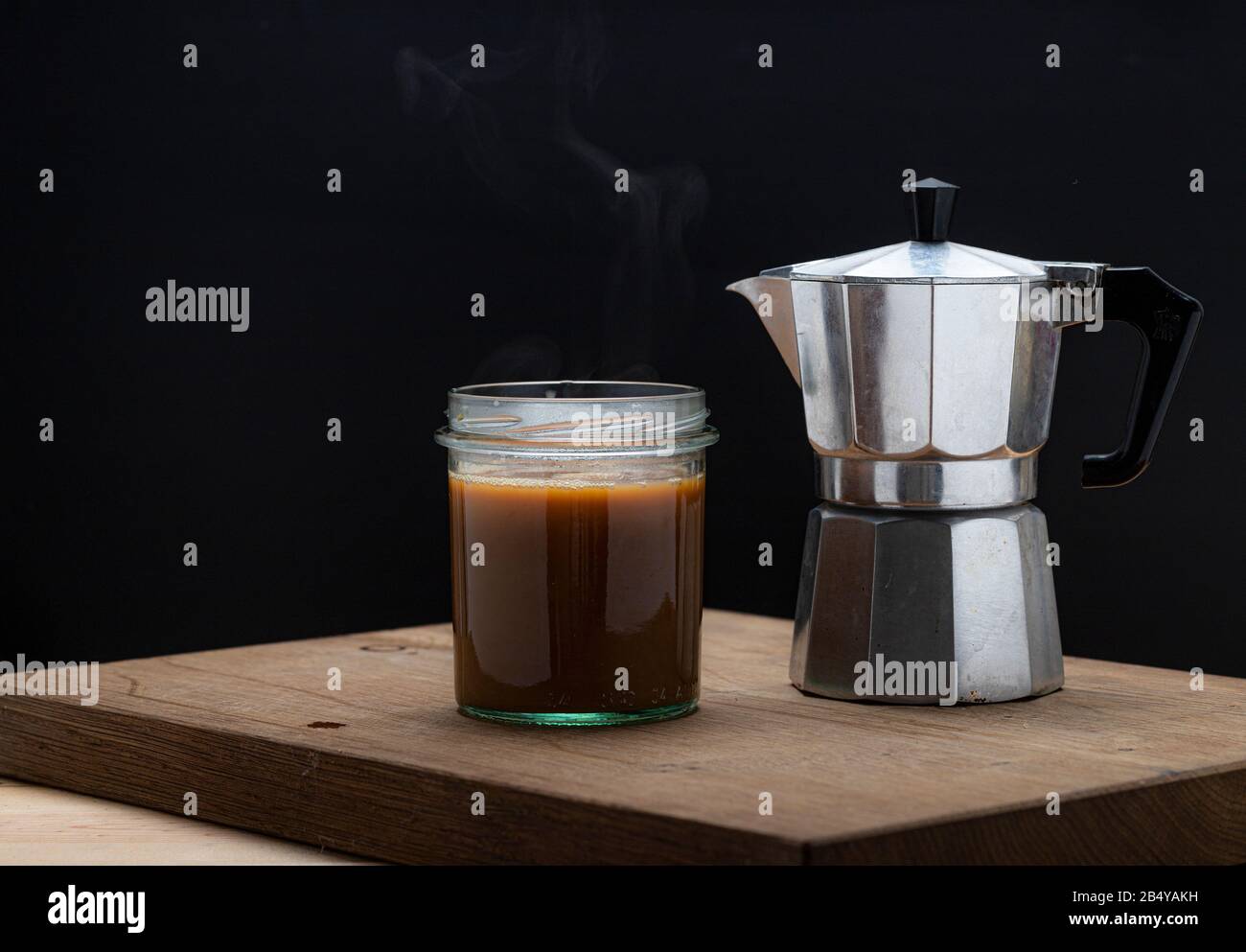 Heißes Dampfgaren erfrischender Kaffee in einem Glas auf dunklem Hintergrund. Gießen von Moka-Hersteller in leeres Glas mit Löffel Stockfoto