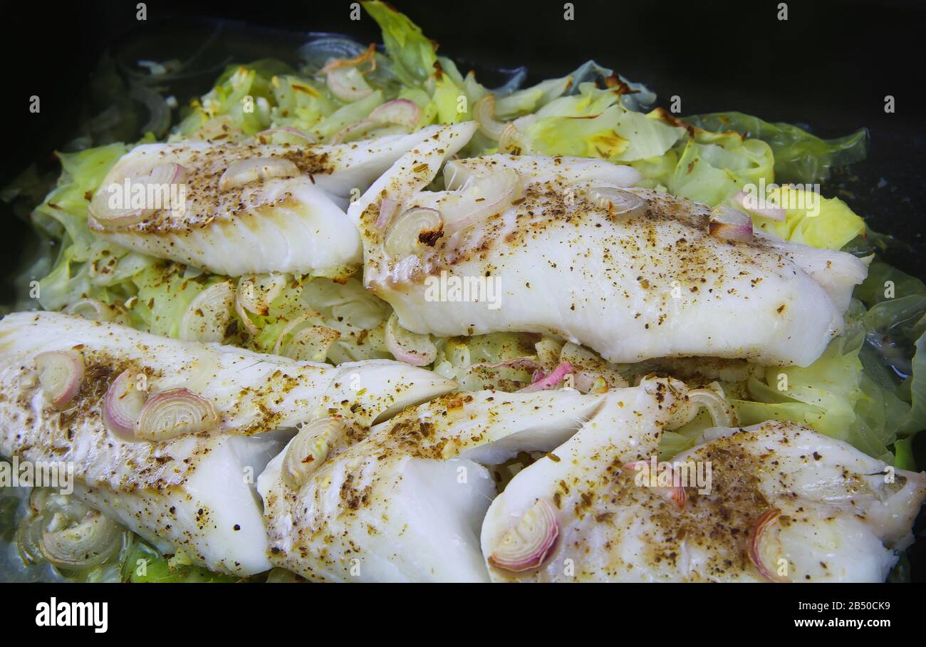Zubereitung der Fischmahlzeit: Blick auf die isolierte Backofentablett mit frischen gekochten Codfisfilets, die auf Spitzkohl gepocht werden, der mit Zwiebeln, Kreuzkümmel und Knoblauch gewürzt ist Stockfoto