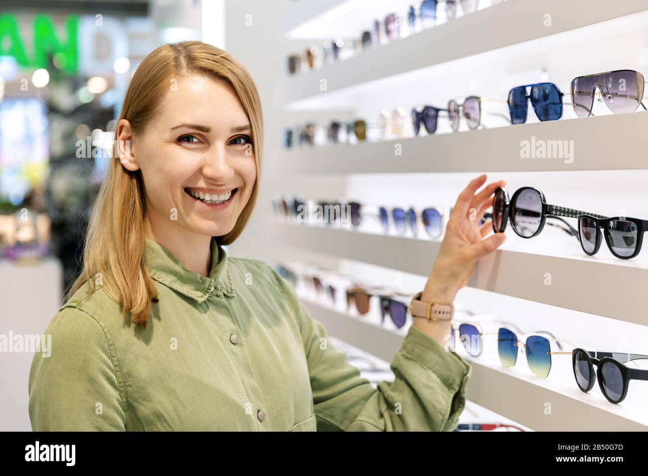 Lächelnde Frau, die sich für eine Sonnenbrille aus dem Brillenregal im Brillenladen entscheidet Stockfoto