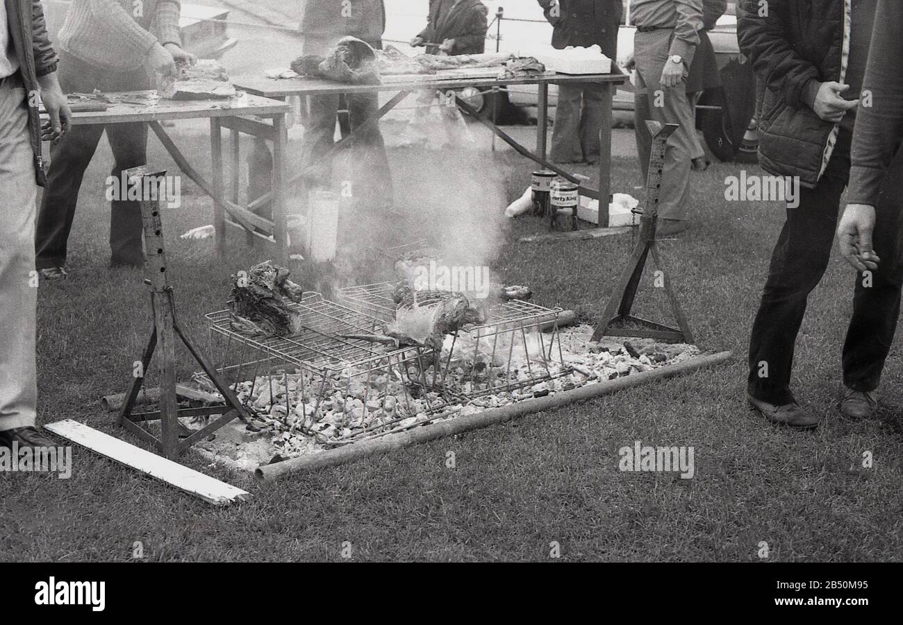 1970er Jahre, historische, große Fleischstücke, die auf einem selbstgebauten Grill zubereitet wurden, ein Drahtgestell über einem offenen Feuer saß auf dem Gras bei einer Veranstaltung im Freien, England, Großbritannien. Stockfoto
