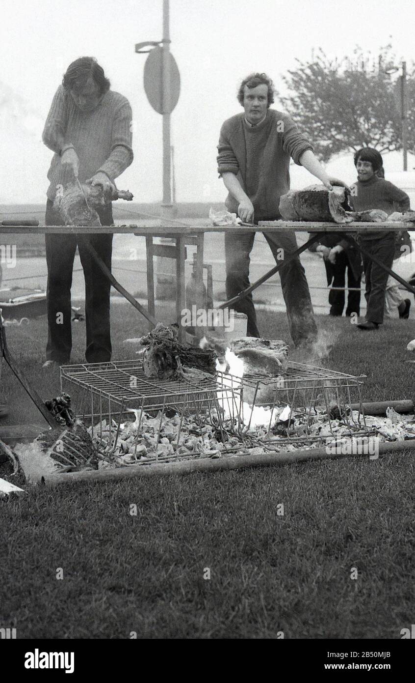1970er Jahre, historische, große Fleischstücke, die auf einem tressel-Tisch geschnitten und auf einem selbstgebauten Grill gekocht wurden, saß ein Drahtgestell über einem offenen Feuer auf dem Gras bei einer Veranstaltung im Freien, England, Großbritannien. Stockfoto