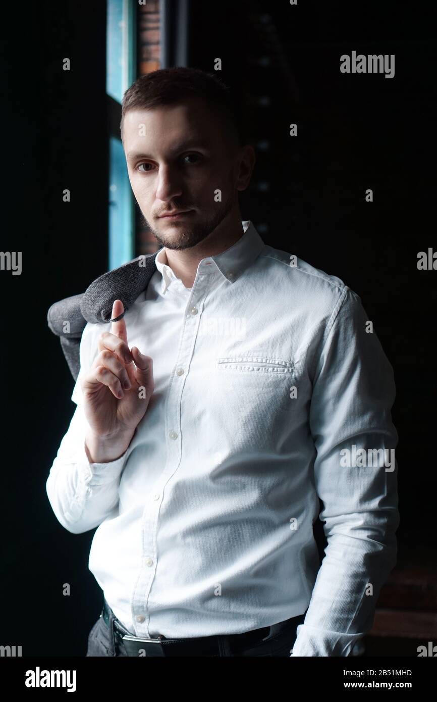 Blick in das Profil eines jungen Geschäftsmannes, gekleidet mit einem weißen Hemd, das in der Nähe des Fensters an einer dunklen Wand steht und die Kamera betrachtet. Vertikales Foto Stockfoto