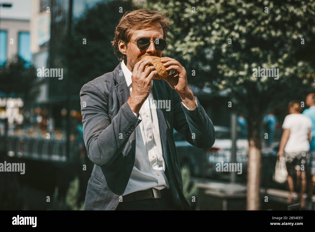 Junger freiberuflicher Geschäftsmann, der einen Hamburger draußen auf einer Straße isst Stockfoto