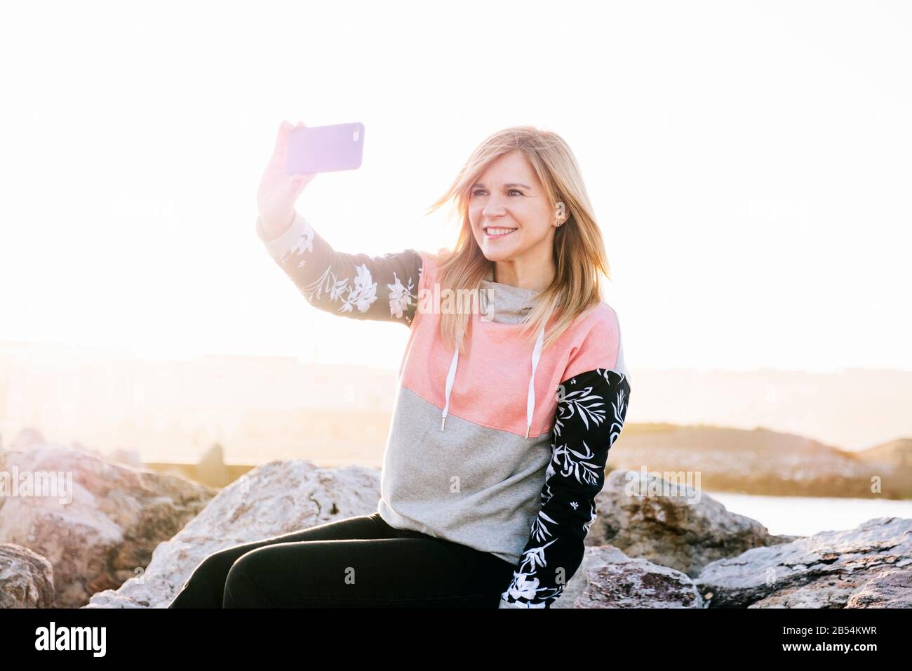 Eine kaukasische blonde Frau mit lässiger Kleidung benutzt das Handy, das an einer Felswand sitzt, für einen Spaziergang am Meer an einem sonnigen Tag bei Sonnenuntergang Stockfoto
