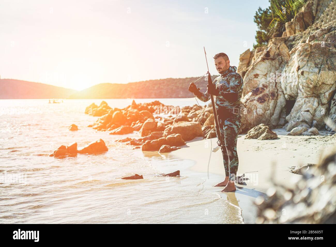 Junger Mann, der in italien beim Sonnenuntergang Speergun für Unterwasserfischen vorbereitet - männlicher Jäger, der sich auf die Jagd vorbereitet, trägt Tarnweatsuit - Extreme s Stockfoto