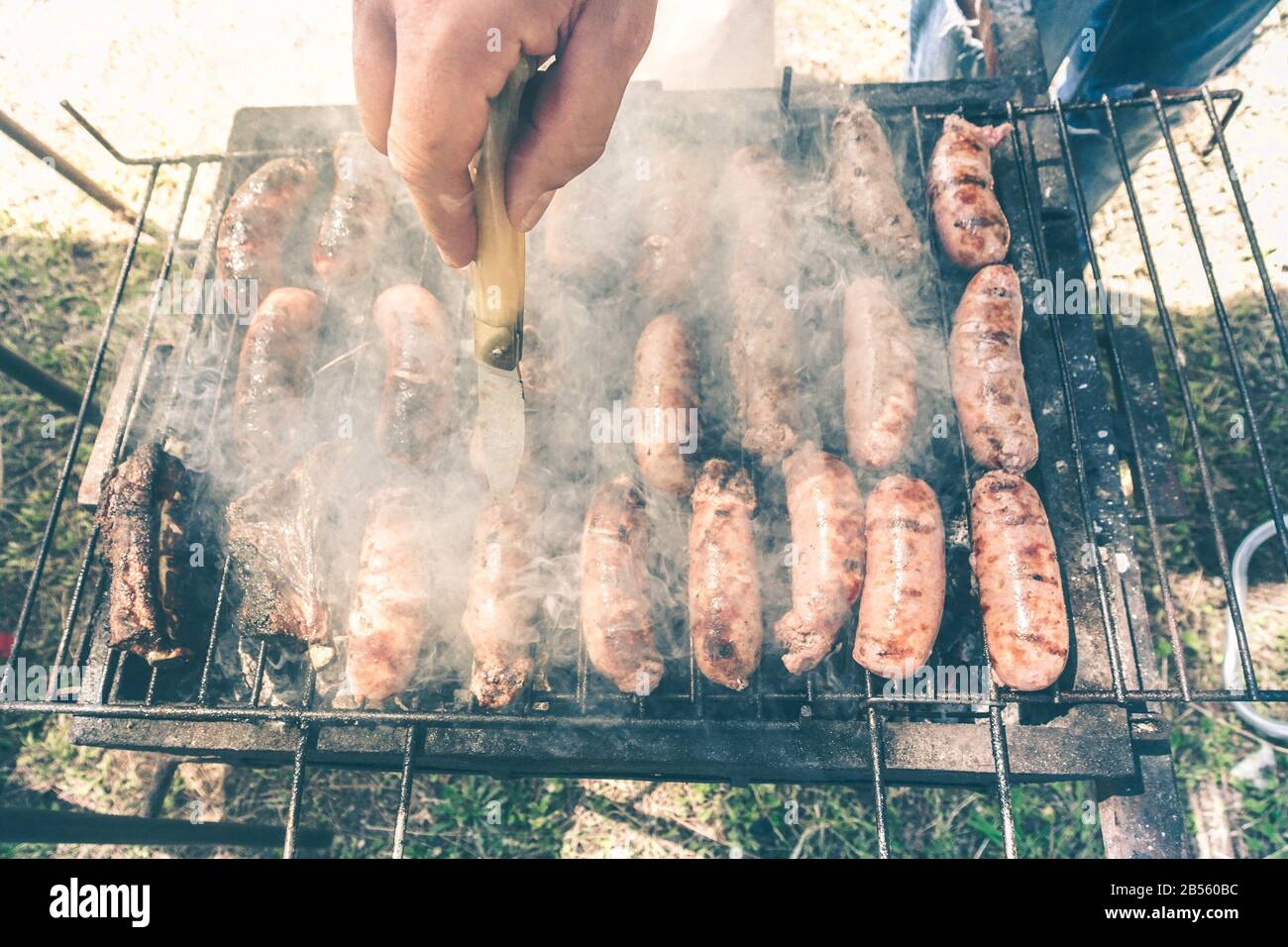 Junger Mann, der Fleisch auf dem Grill kocht - Chef, der einige Schweinefleischwürste im Freien im Park auf den Grill legt - Konzept des Essens von bbq im Freien während der Sommerzeit - Sof Stockfoto