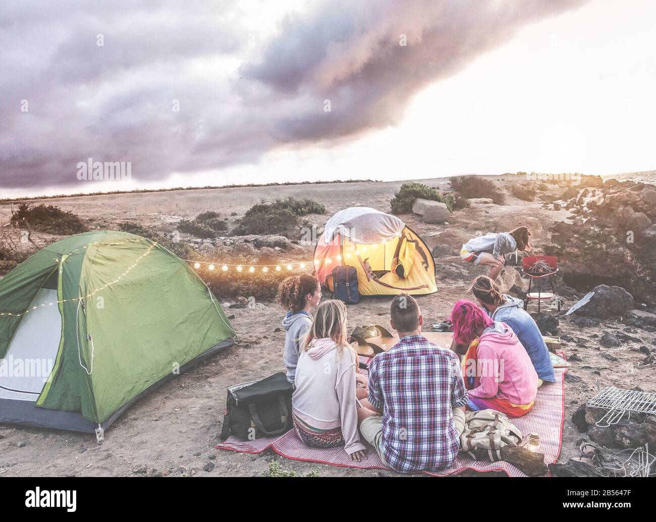 Fröhliche Freunde, die bei Sonnenuntergang in der Wüste ein Picknick machen - Junge Leute haben Spaß beim Abendessen - Reisen, Abenteuer auf Tour in der Natur, vac Stockfoto