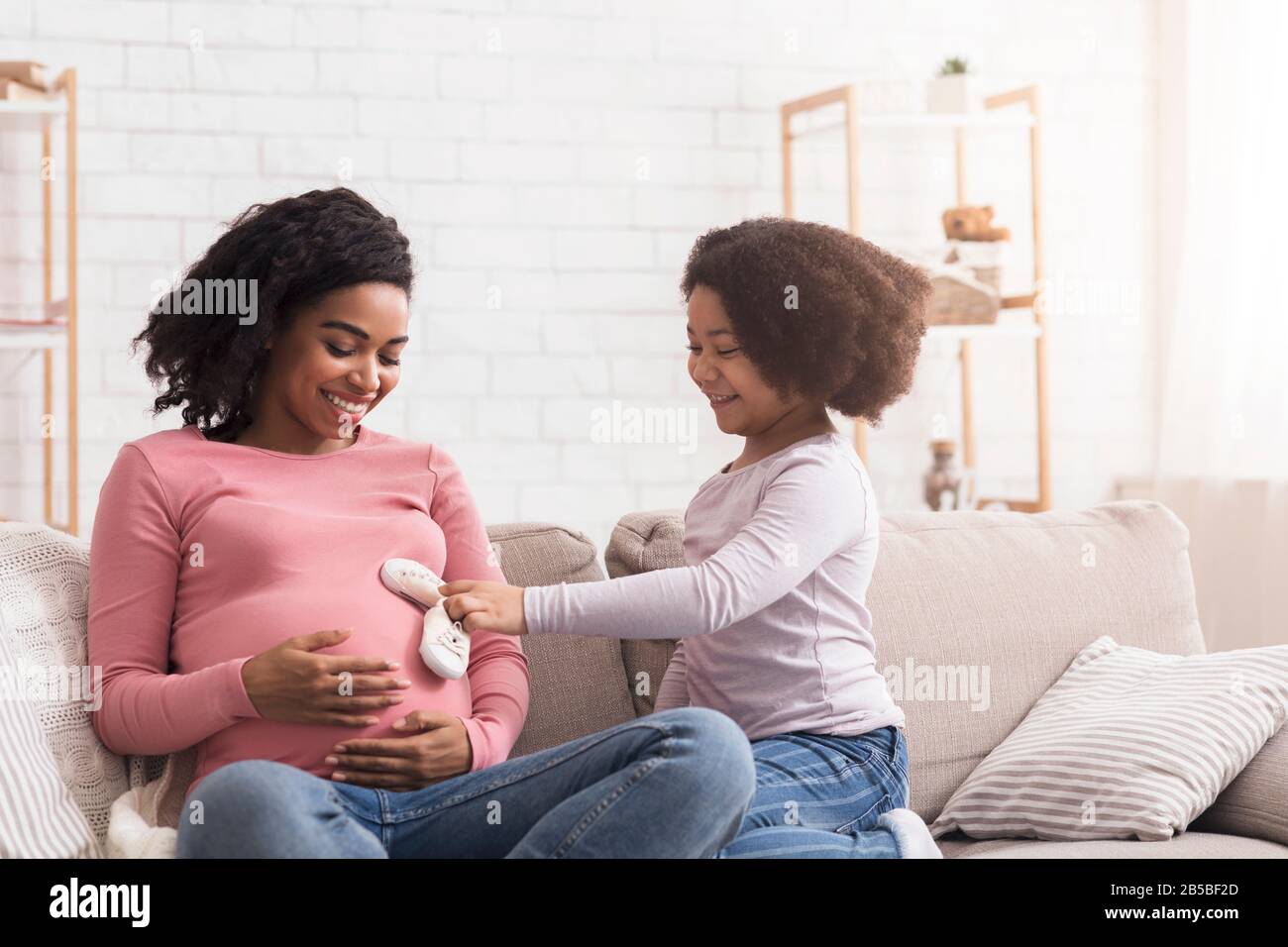 Verspieltes kleines afro-Mädchen, das Babyschuhe an den Bauch der schwangeren Mutter legt Stockfoto