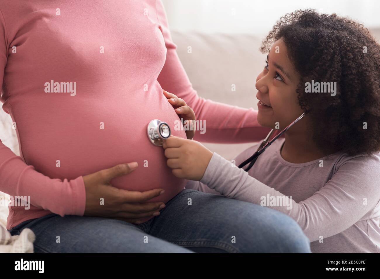Kleine afro-Tochter, die mit Stethoskop dem Bauch ihrer schwangeren Mutter lauscht Stockfoto