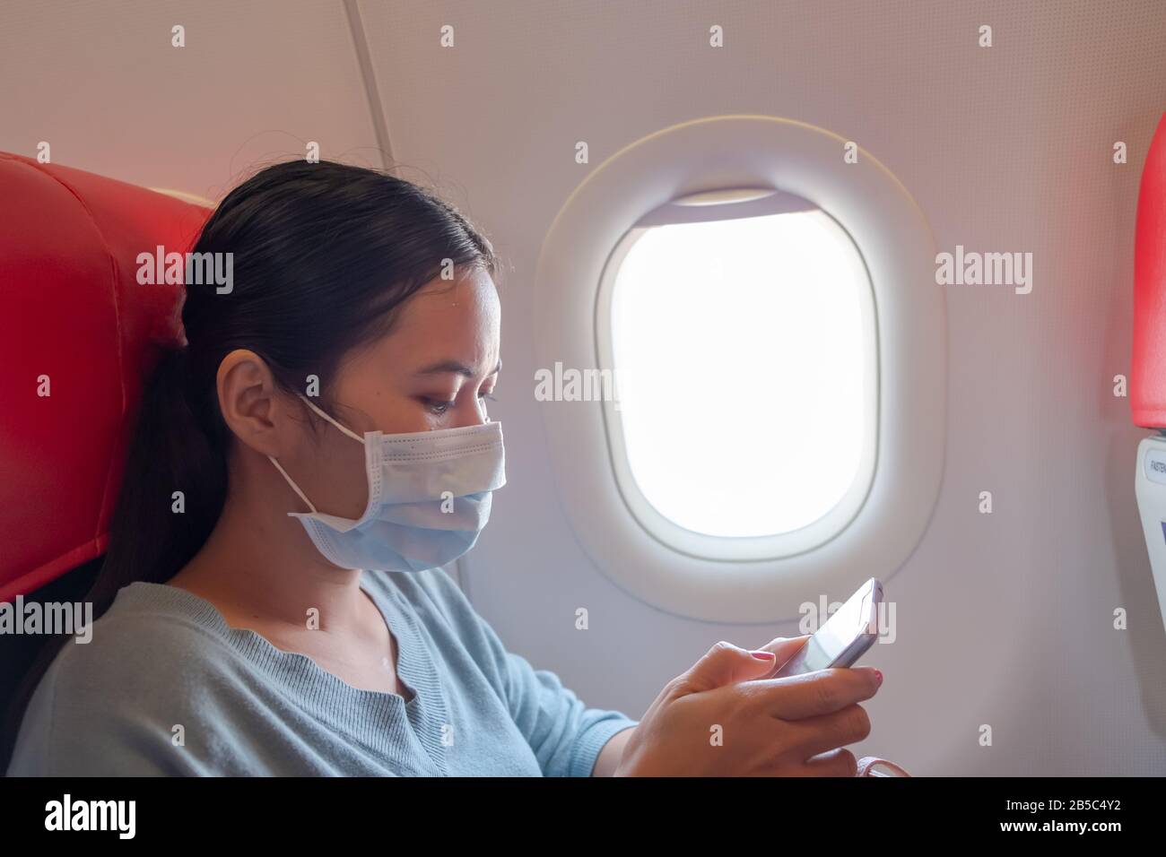 Asiatische Mädchen verwenden eine Schutzmaske für Coronavirus oder Kovid 19 im Flughafen. Thailändische Dame, die mit ihrem Telefon im Flugzeug spielt. Stockfoto