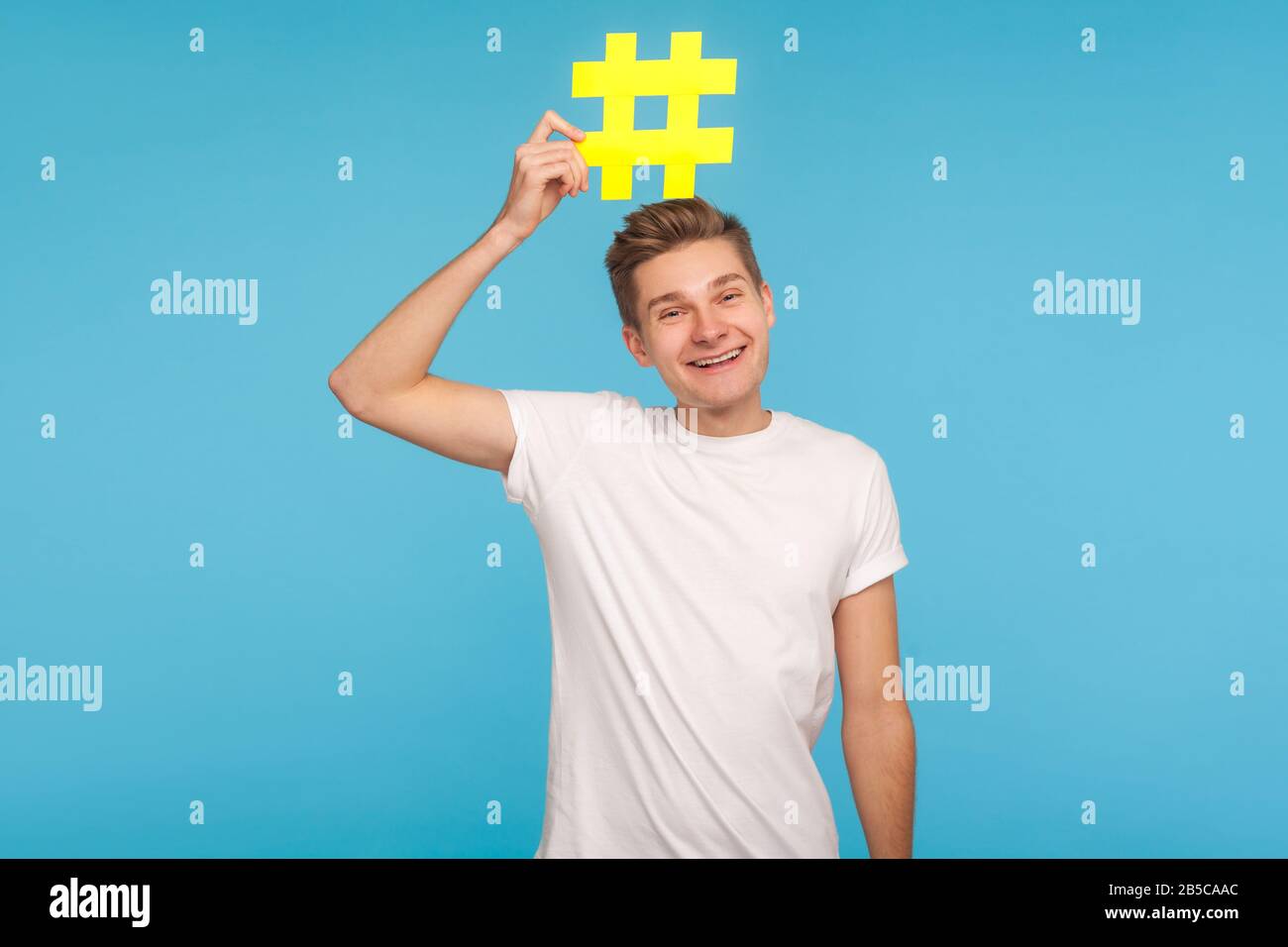 Medienpopularität. Unbeschwerter lustiger Mann im T-Shirt, der großes gelbes Hashtag-Symbol auf dem Kopf hält und lächelt, Hash-Zeichen von Internettrends, virales soziales Stockfoto
