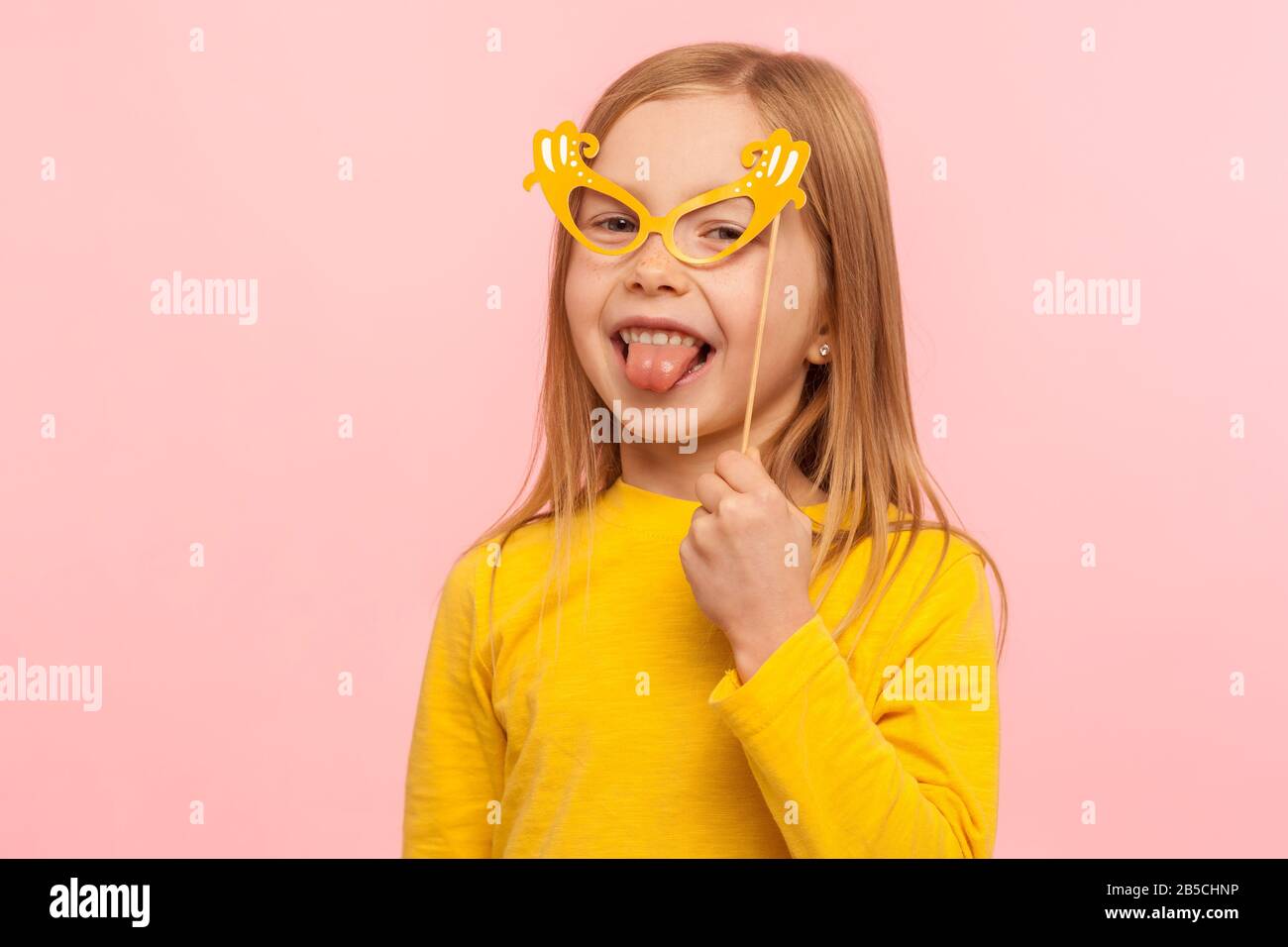 Portrait des charmanten Ingwer-Mädchens, das die Augen mit Papierbrille bedeckt und die Zunge herausklebt, frech Kind hat Spaß und trägt Maskenmaske. I Stockfoto