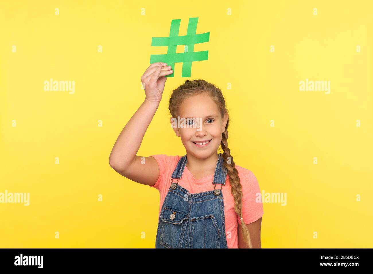 Virale Webinhalte, Internetforum. Porträt des glücklichen kleinen Mädchens mit Geflecht in Denim-Overalls, das Hashtag-Symbol über Kopf hält und lächelt, wirbt Stockfoto