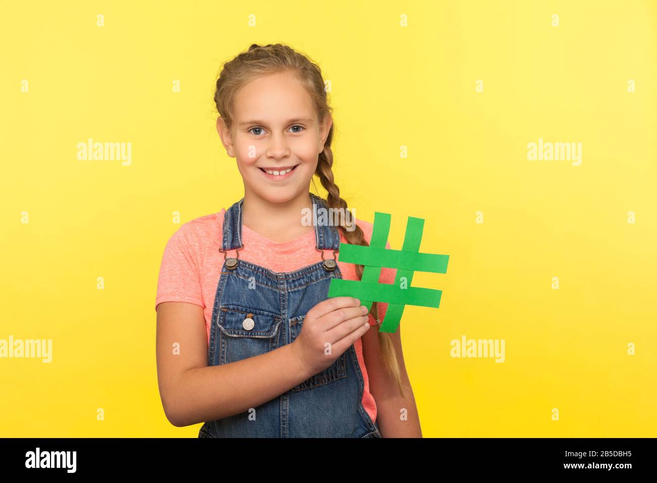 Internettrends, virale Webinhalte. Portrait des fröhlichen kleinen Mädchens mit Geflecht in Denim-Overalls, das Hashtag-Symbol und lächelnd, Werbeschild hält Stockfoto