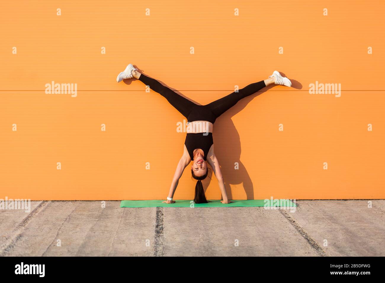 Überfreuliches glückliches Mädchen mit perfektem sportlichem Körper in enger Sportswear, das Yoga-Stand-Pose mit gespreizten Beinen gegen die Wand macht und Zunge zeigt, habend Stockfoto