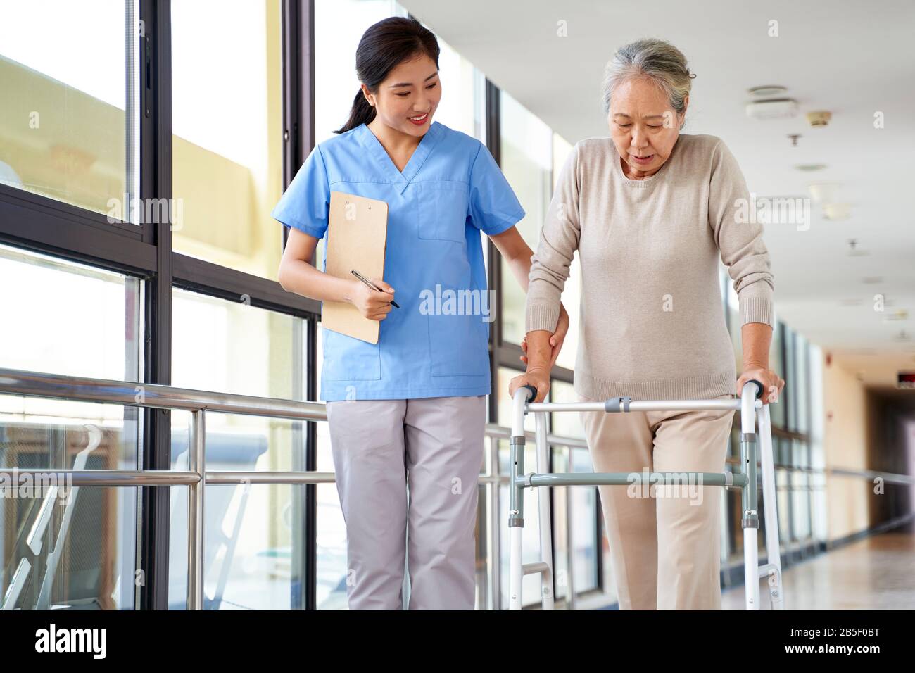Junger asiatischer Physiotherapeut, der mit einer älteren Frau beim gehen mit einem Wanderer arbeitet Stockfoto