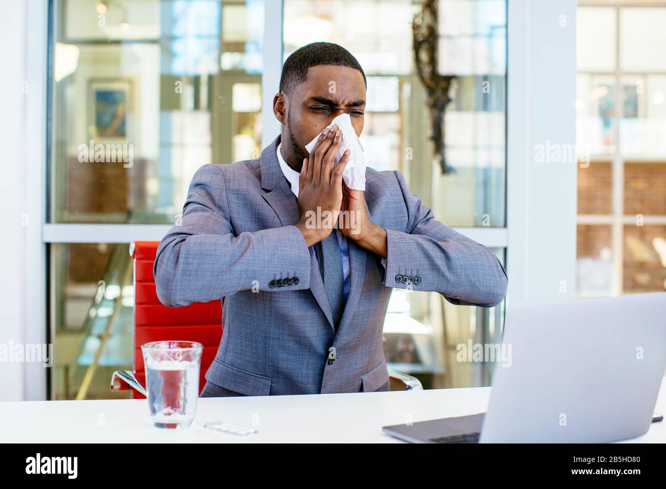 Porträt eines kranken jungen Mannes im Business-Anzug, der seine Nase weht, während er hinter dem Schreibtisch bei der Arbeit mit dem Computer sitzt Stockfoto