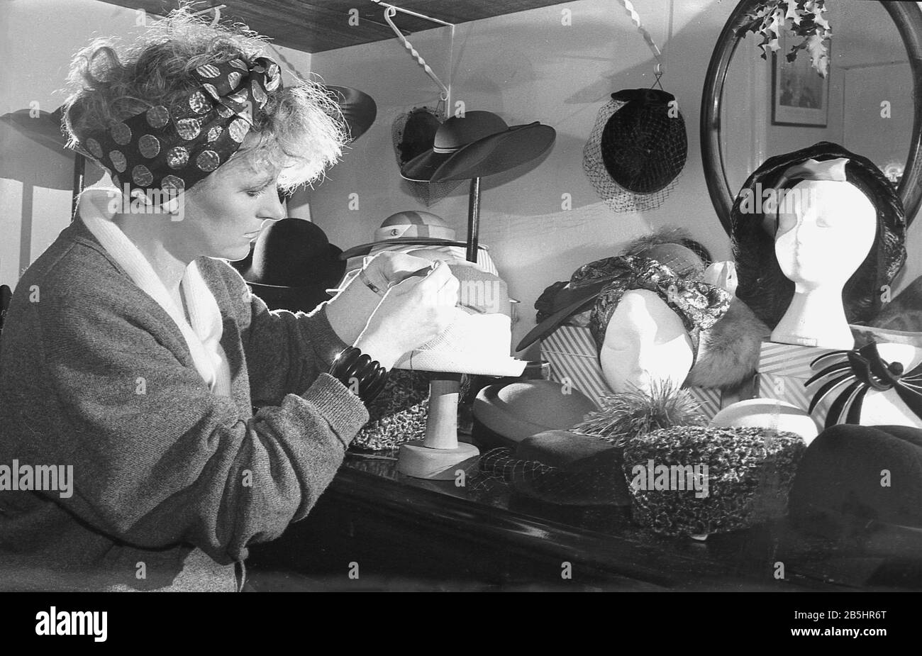 In den 1980er Jahren zeigt das Foto eine Hutmacherin oder Mühlerin, die in ihrem Handwerk sitzt, mit einer Tischausstellung ihrer Kopfbekleidungskreationen in England, Großbritannien. Die Mühlerei wird oft von unabhängigen Arbeitern betrieben, Einzelhändlern, die die Design- und kreativen Aspekte der maßgeschneiderten Hutmacherei genießen. Stockfoto