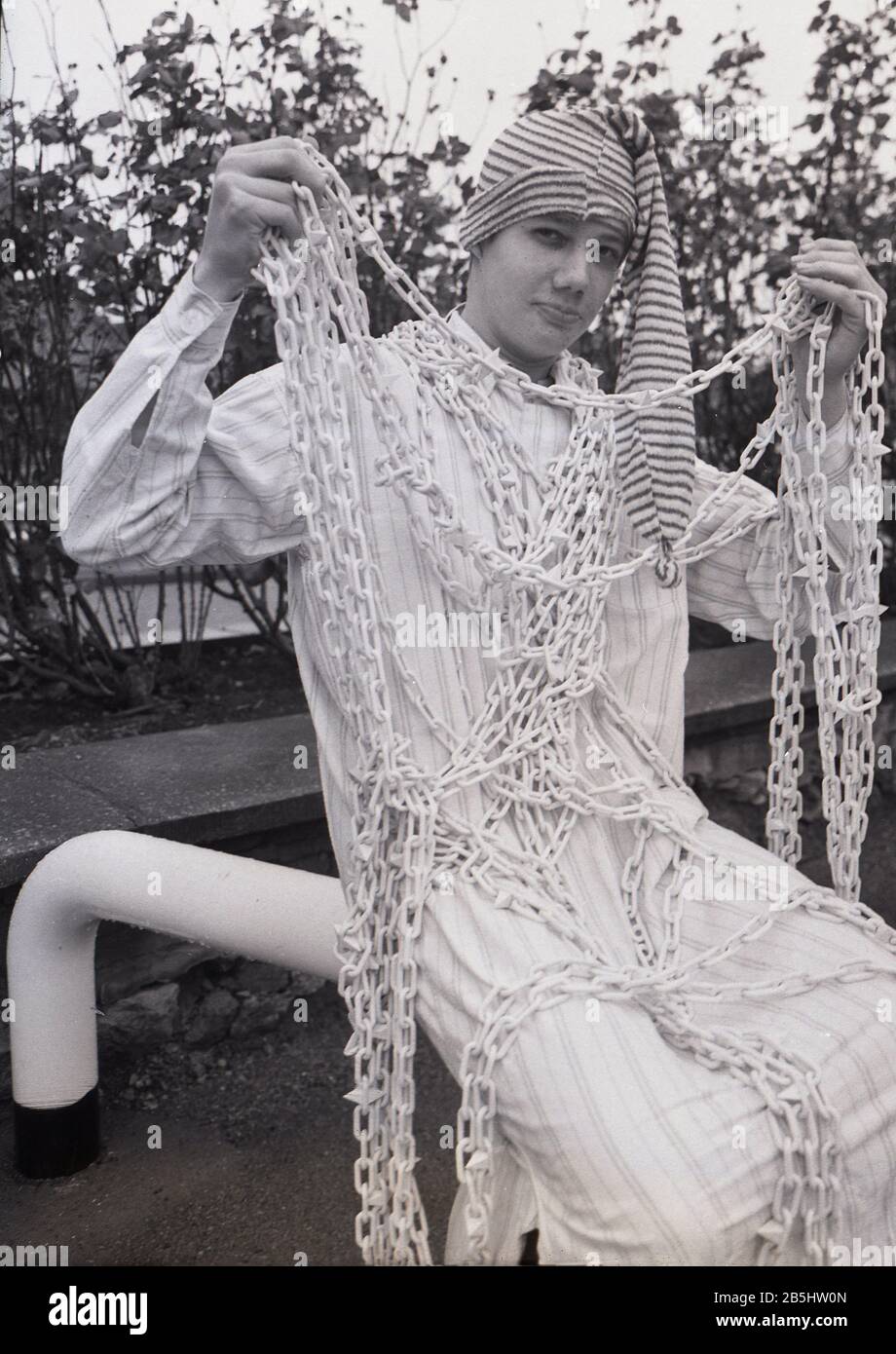 1980er Jahre, historischer junger Mann, der sich als Figur aus dem Roman „Silas Marner“ verkleidet hat, der Geist oder übernatürlich bei einem carol-Gesangsevent in England, Großbritannien, mit einem knöchellangen Nachthemd aus Baumwolle, einem winzigen Hut und einer Stoffkette. Stockfoto