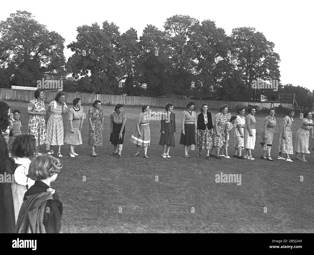 1950er Jahre, historisch, beobachtet von lokalen Kindern, einer Gruppe von Damen, Müttern und Hausfrauen, die die langen Kleider der Ära mit Blumenmuster tragen, auf einem Feld aneinandergereiht sind, bereit für ein kurzes Laufrennen oder Sprint, als Teil eines Community-Spaßtages, England, Großbritannien. Stockfoto