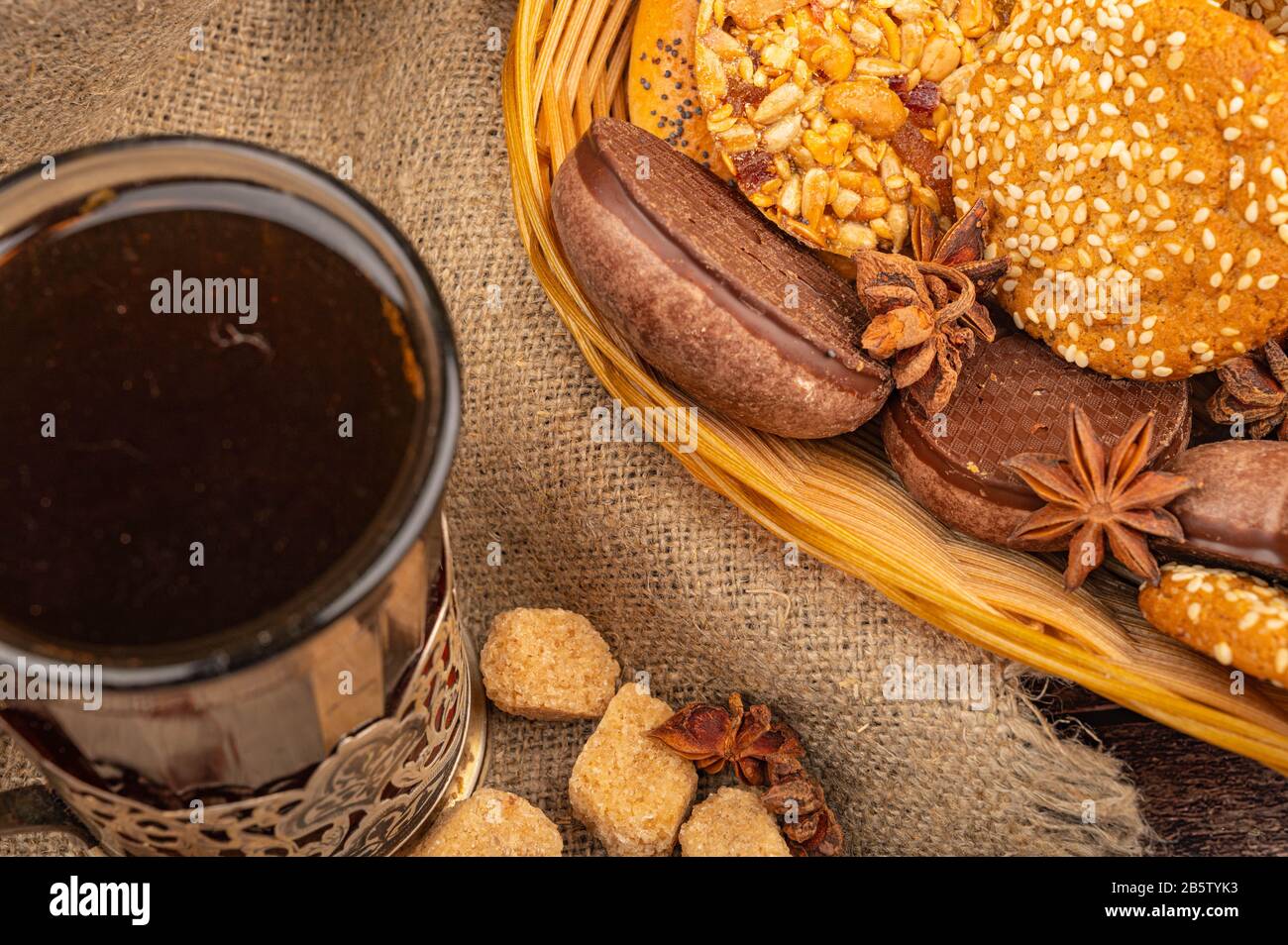 Ein Glas Tee in einem Vintage-Cup-Halter und Plätzchen, Schokoladenkuchen und Sternanis in einem Korbkorb auf dem Hintergrund rauer Homespun-Qualität. Schließen Stockfoto