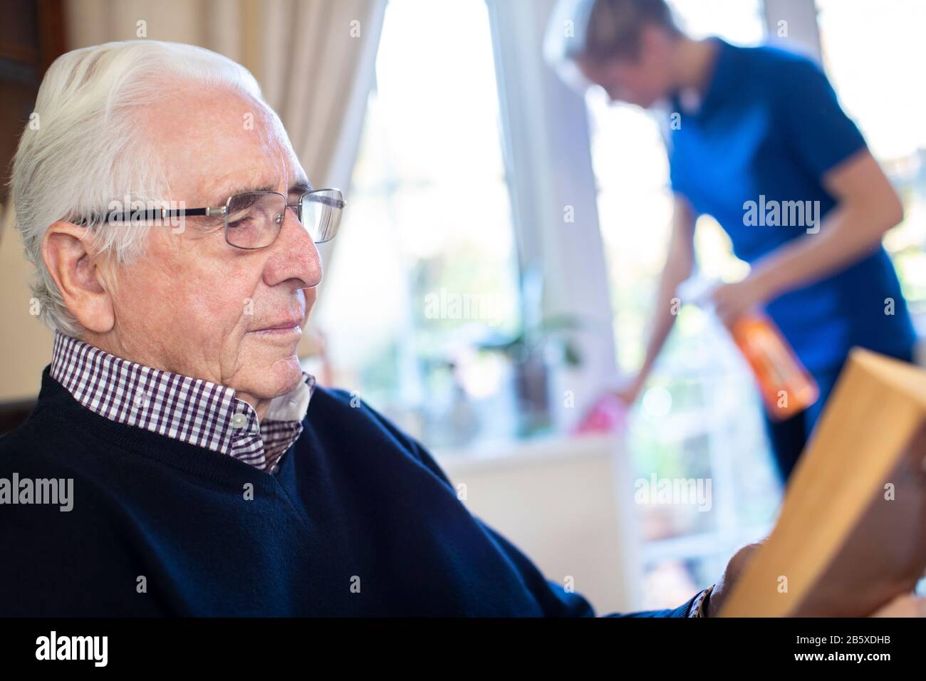Der Ältere Mann Liest Im Vordergrund, Während Die Weibliche Hilfe Das Haus Reinigt Stockfoto