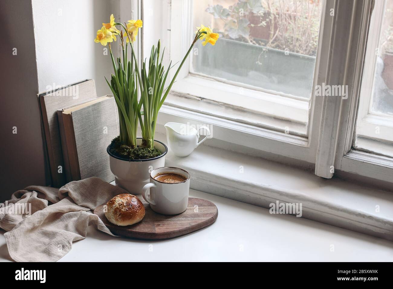 Gemütliches Stilllebensszenerie im Osterfrühling. Tasse Kaffee, süßer Brötchen, Bücher, Holzschneidebrett, Milchkrug und Blumenvase auf altem Fensterbrett. Blumen Stockfoto