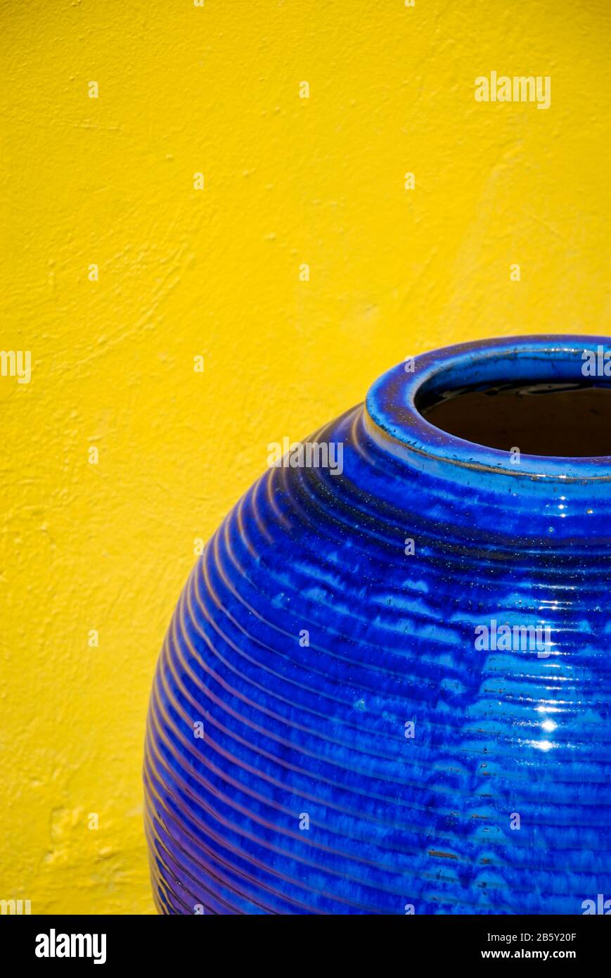 Große, glänzende, strukturierte blaue Vase, die im Freien neben einem hellen tropisch-gelben Wandkopierraum sitzt Stockfoto