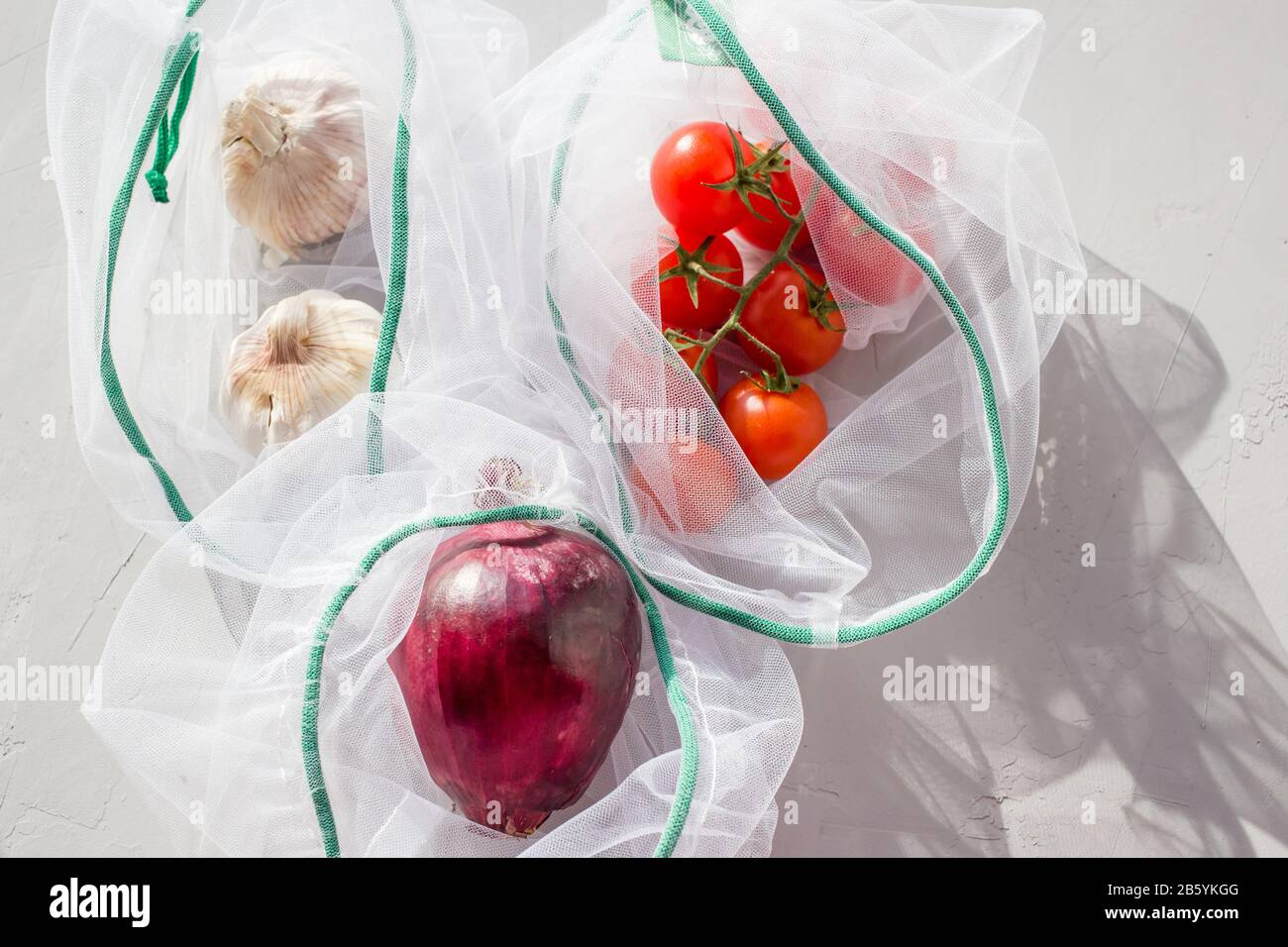 Die wiederverwendbare Netztasche aus Organza zum Einkaufen mit Tomaten, Zwiebel und Knoblauch auf grauem Hintergrund. Konzept: Kein Kunststoff, kein Abfall, wiederverwendbare Lebensdauer. Stockfoto