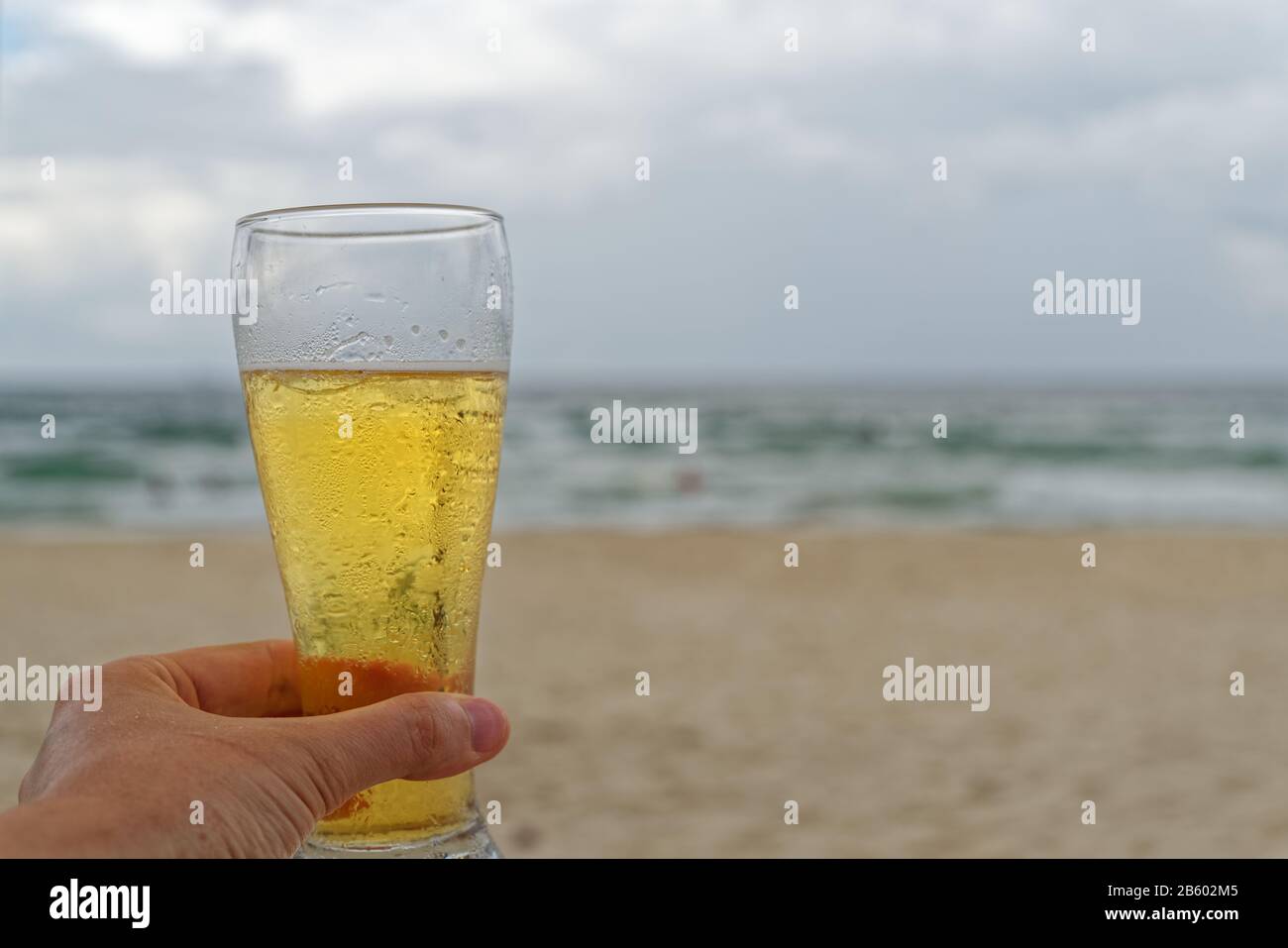 Männliche Hand hält ein Glas Bier am Strand vor bewölktem Himmel Stockfoto