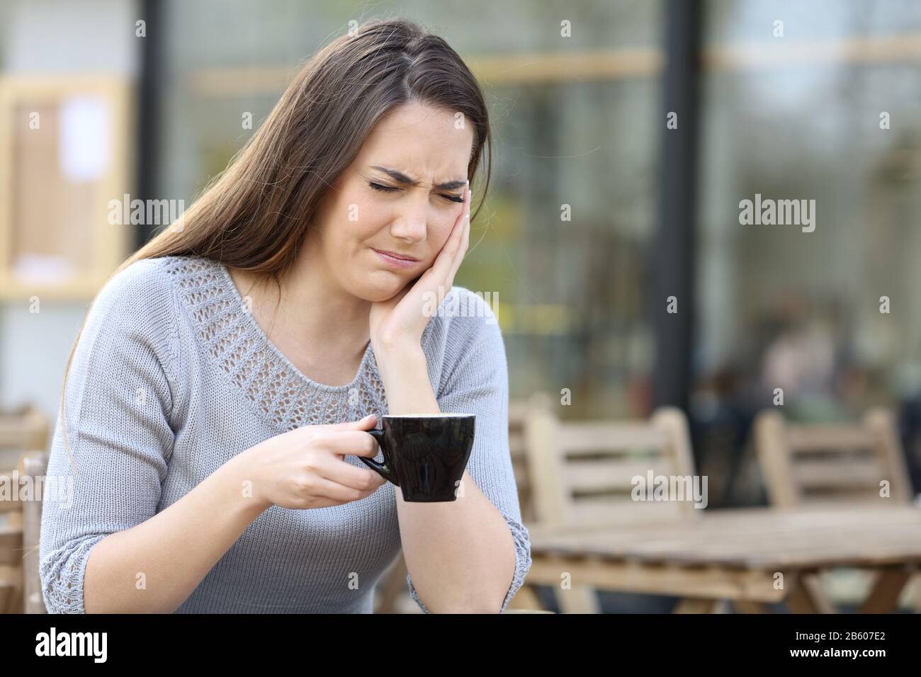 Frau, die an schmerzhaften Zahnschmerzen leidet und einen Kaffeebecher auf einer Restaurantterrasse hält Stockfoto