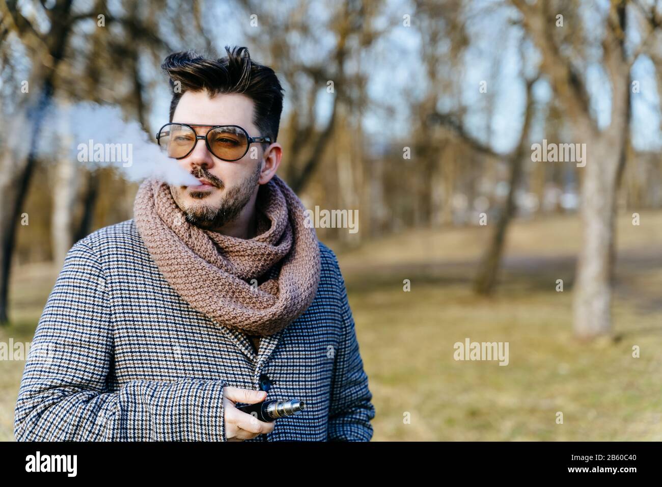 Selbstbewusster junger bärtiger Mann in Sonnenbrille schäumt elektronische Zigarette. Ein junger, hübsch bärtiger Flusspferd, der im Park eine E-Zigarette im Freien umgibt Stockfoto
