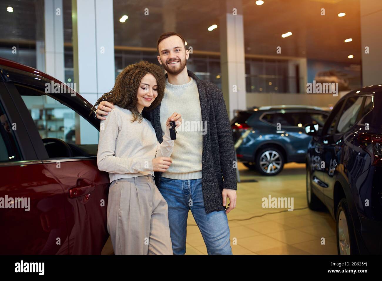 Gut aussehendes, fröhliches Mädchen mit gewelltem, lockigem Harir, das an den Schlüsseln des luxuriösen roten Autos liegt, Mann umarmt, seine Frau umarmt, Nahaufnahme Foto. Mann präsentiert Stockfoto
