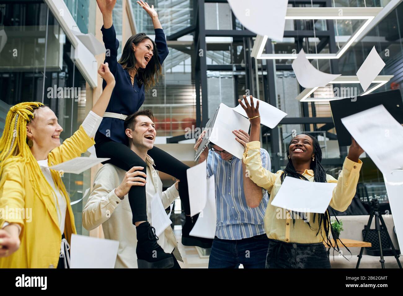 Kreativ freundliche Mitarbeiter, die im Büro hängen, Angestellte, die Dokumente, Papiere, schreiende, brünette Frau werfen, die auf den Scholidern ihres c sitzt Stockfoto