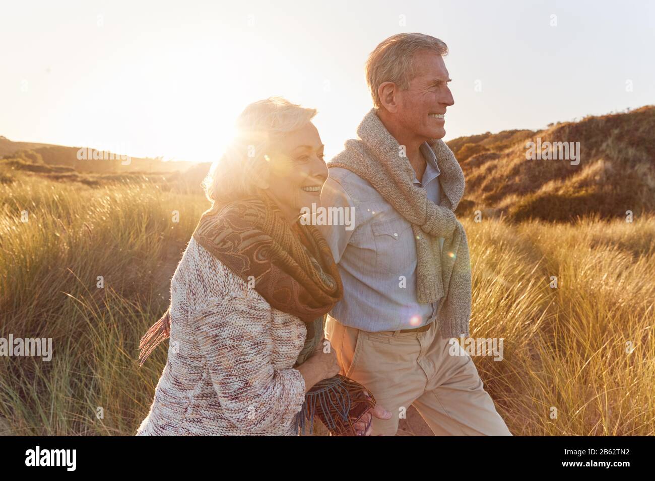 Liebende Pensionierte Paare, Die Arm In Arm Durch Sanddünen Im Strandurlaub Gegen Die Aufflackernde Sonne Laufen Stockfoto