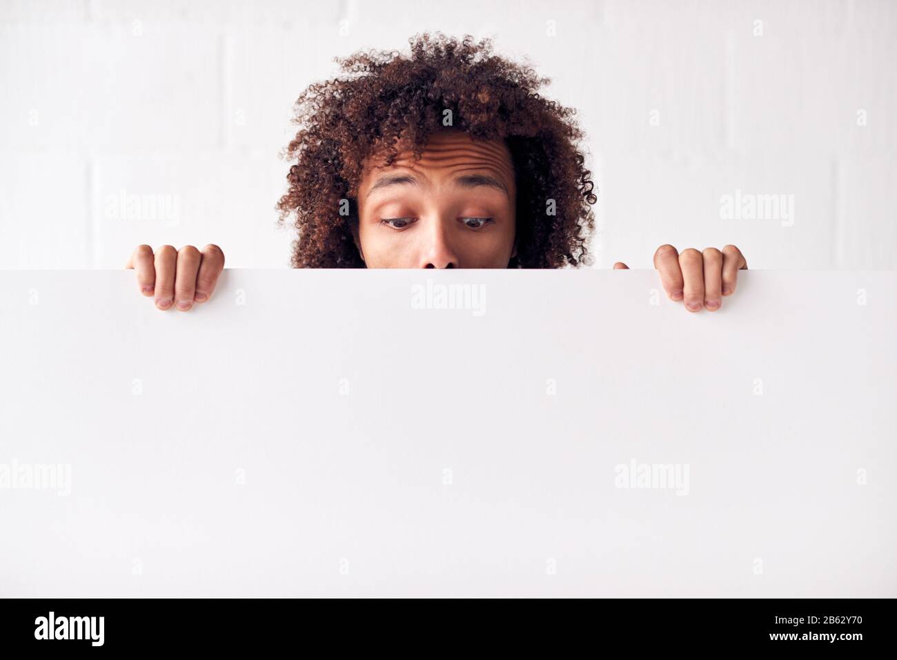 Happy Young Man Hält Leere Weiße Karte Mit Copy Space, Der Gegen Die Wand Des Weißen Studios Steht Stockfoto
