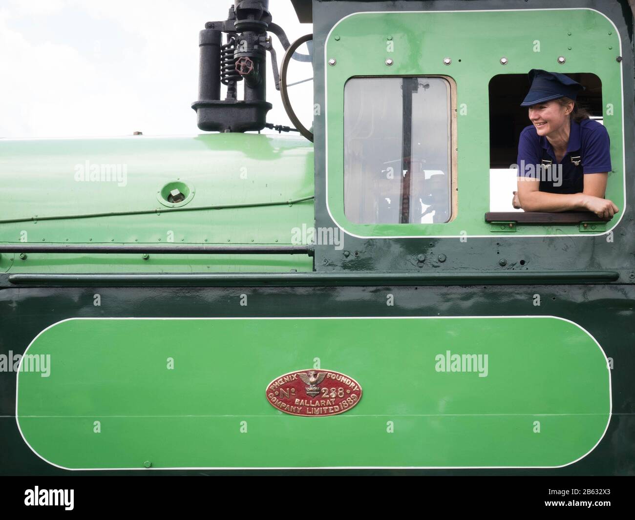 Junge Frauen Steamrail Victoria Freiwillige Dampflok-Ingenieur lehnt sich grün und schwarz Lok-Motorfenster lächelnd an sonnigen Tag. Stockfoto
