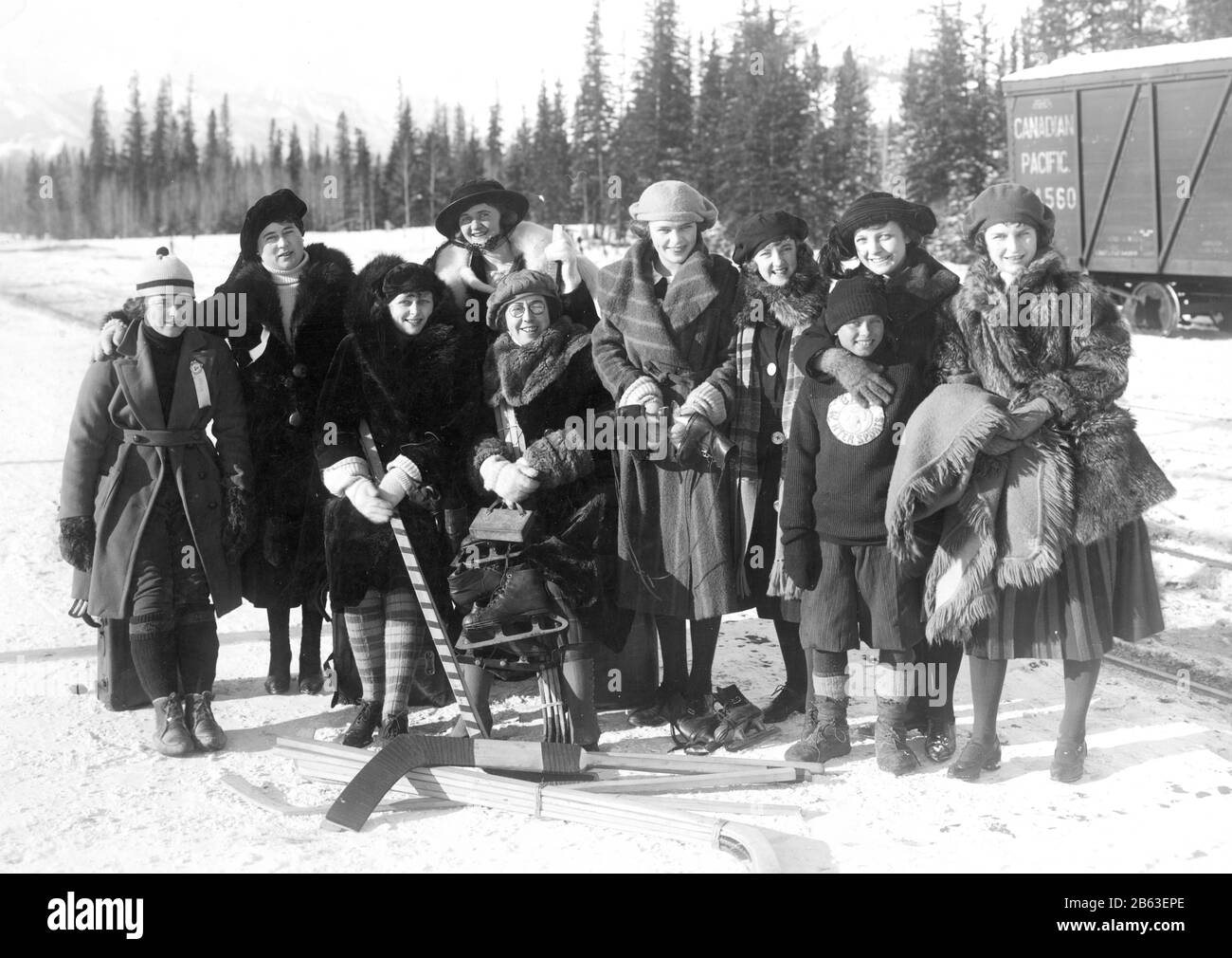 Frauen und Mädchen Hockeyspieler und -Familie stehen etwa 1920 neben Bahngleisen in Calgary, Kanada. Sie posieren mit Hockeyschlägern, Schlittschuhen usw. und tragen schwere Winterkleidung und sehen alle sehr glücklich aus. Ein Mädchen auf der linken Seite trägt ein Band, das sagt: "Offiziell". Ein Junge auf der rechten Seite hat einen gut getragenen Flecken, der sagt: "Wintersport", um meine zugehörigen Vintage-Bilder zu sehen, Suche: Prestor Vintage Sport Woman Stockfoto
