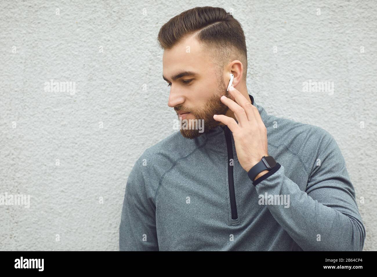 Bärtiger Kerl in grauer Sportswear mit Kopfhörern auf grauem Hintergrund. Stockfoto